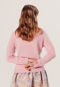Rosa gestrickter Pullover mit langen Ärmeln und gerippten Bündchen, getragen über einem gemusterten Blumenrock, von hinten betrachtet.