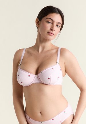 Kobieta ubrana w jasnoróżowy biustonosz i pasujące majtki z małymi czerwonymi nadrukami wiśni, stojąca na neutralnym tle.