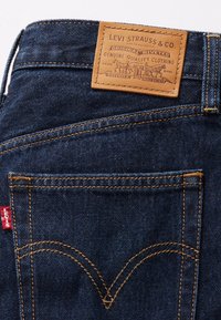 Jean en denim bleu foncé avec un patch de marque en cuir marron, des coutures dorées visibles et une étiquette rouge près de la poche arrière avec un design signature.