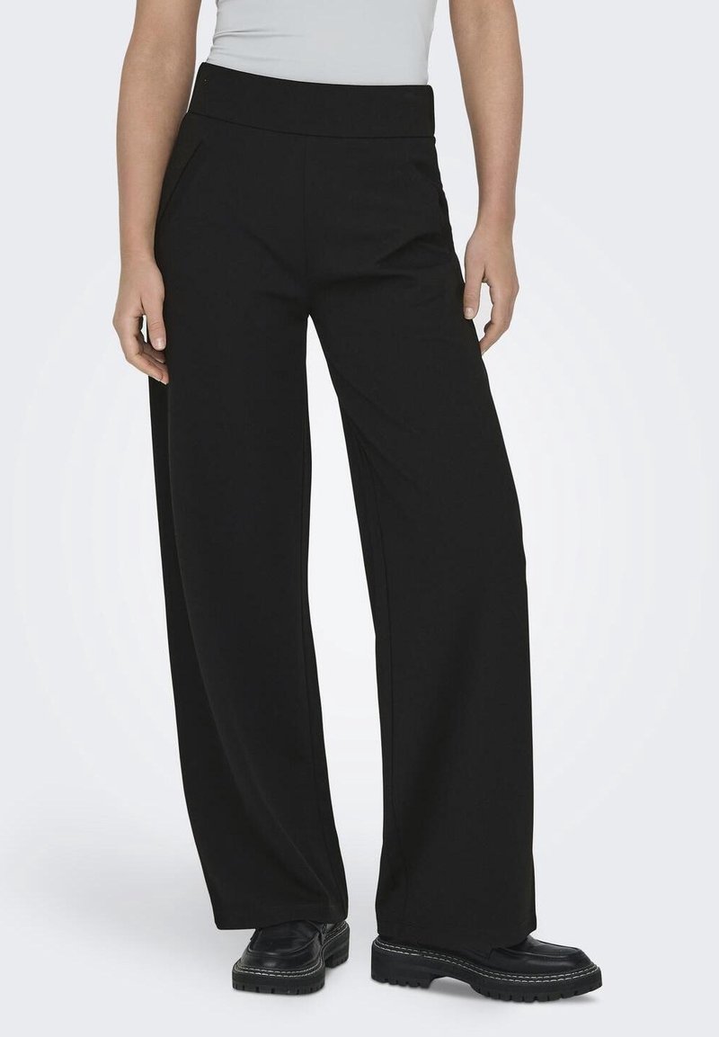 Femme portant un pantalon noir à jambes larges, des chaussures noires épaisses et un haut gris clair rentré, debout devant un fond uni.