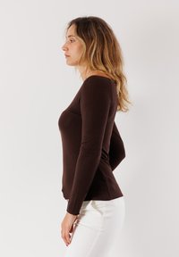 Maglietta marrone a maniche lunghe realizzata in tessuto elasticizzato, forma attillata, scollatura rotonda e texture liscia, abbinata a jeans bianchi.