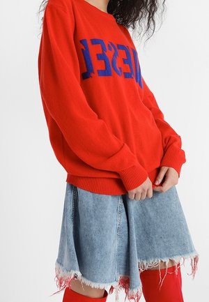 Persoon draagt een rode trui met blauwe tekst, een versleten lichte spijkerrok en rode over-de-knie laarzen tegen een effen witte achtergrond.