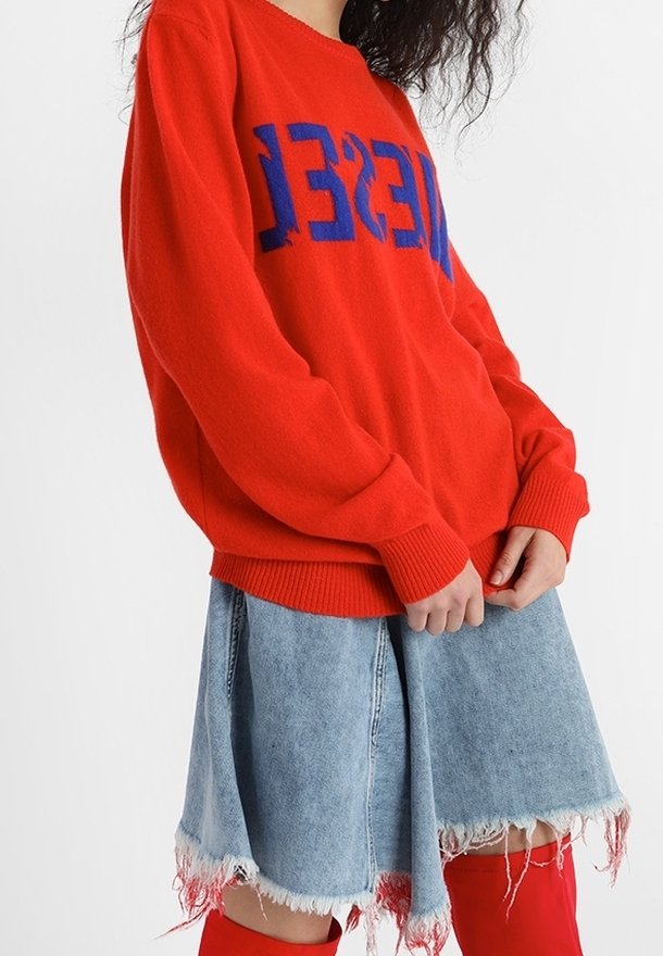 Personne portant un pull rouge avec un texte bleu, une jupe en jean clair effilochée et des bottes rouges montantes au-dessus des genoux, sur un fond blanc uni.