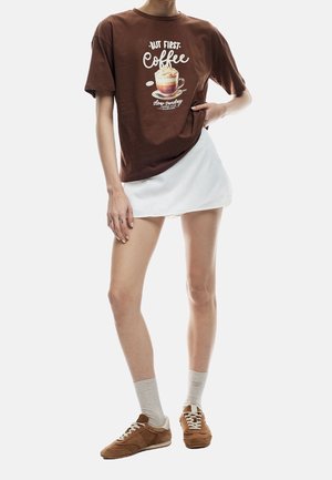Osoba ubrana w brązowy t-shirt z grafiką kawy, białą mini spódnicę, jasne skarpetki oraz brązowe sneakersy, stojąca na jednolitym tle.