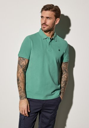 Polo shirt verde con colletto, maniche corte e un piccolo logo sul petto, abbinato a pantaloni cargo scuri di colore navy e avambracci tatuati.