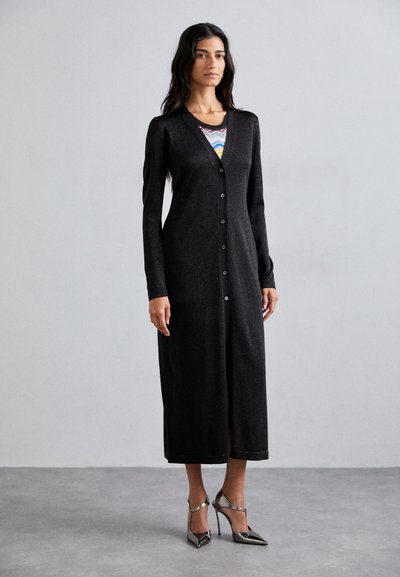 Long cardigan noir en tissu doux et texturé, avec un col en V, une fermeture à boutons et des manches longues, associé à des talons hauts argentés.