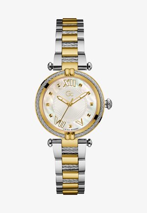 Zelta un sudraba toņu pulkstenis ar apaļu mātes pērļu ciparnīcu, kas apzīmēta ar romiešu cipariem, un teksturētu divu toņu metāla siksnu.