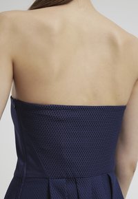Robe sans bretelles de couleur marine avec un motif texturé en damier. Présente un corsage ajusté, une fermeture éclair dissimulée sur le côté et une jupe évasée.