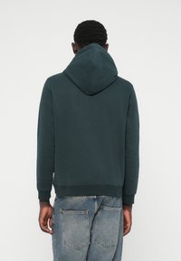 Sweatshirt à capuche vert foncé avec une coupe décontractée, des poignets et un ourlet côtelés, comportant une poche kangourou et un tissu doux et texturé.