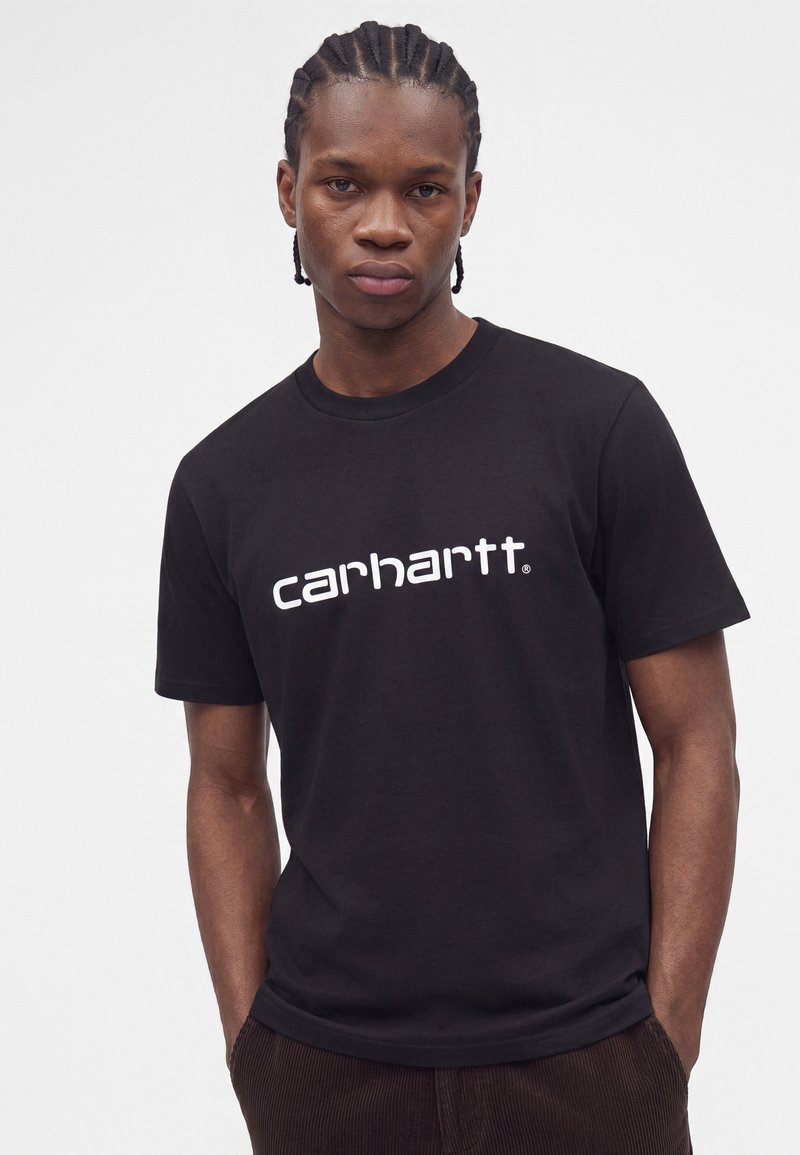 Junger Mann mit geflochtenem Haar, der ein schwarzes Carhartt T-Shirt und dunkle Hose vor einem schlichten hellen Hintergrund trägt.