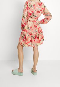 Žena v dlouhé růžové květinové šatech s dlouhým rukávem a světle zelených platformových pantoflích, stojící s rukou na boku, obrácená zády.