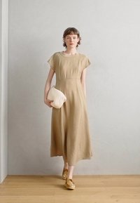 Beige midi dress with short sleeves, round neckline, and a flowy silhouette. Model holds a soft, cream-colored pouch. Wooden floor backdrop.