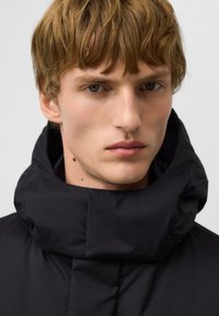 Czarna kurtka puchowa z wysokim kołnierzem, o gładkiej teksturze i minimalistycznym wzornictwie. Model ma jasne włosy i neutralne rysy twarzy.