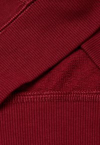 Roter Stoff mit zwei überlappenden Abschnitten: ein gerippter und ein weiterer mit einem strukturierten Muster. Sichtbare Nähte entlang der Kanten.