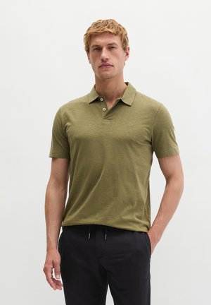 Mężczyzna w oliwkowo-zielonej koszulce polo i czarnych spodniach, stoi z jedną ręką w kieszeni na jednolitym tle.