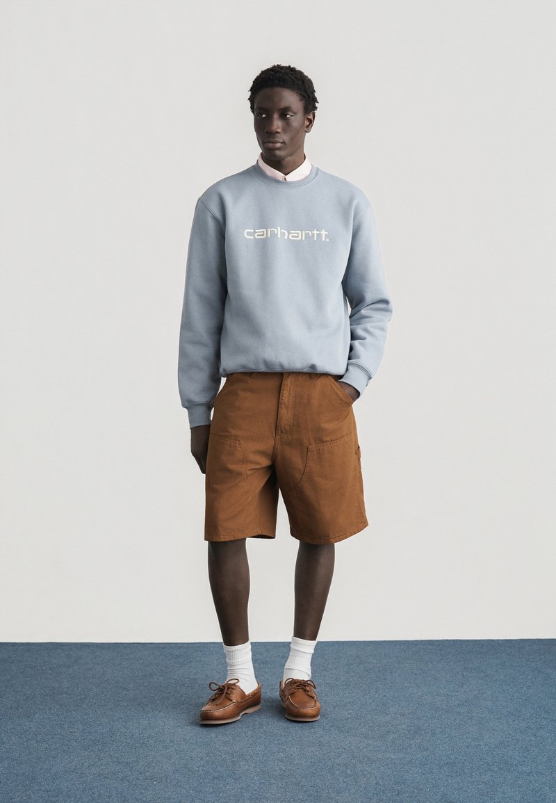 Junger Mann steht vor schlichtem Hintergrund und trägt ein hellblaues Carhartt-Sweatshirt, braune kniehohe Shorts, weiße Socken und braune Lederschuhe.