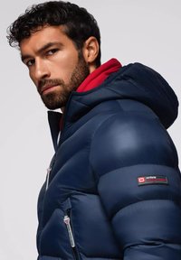 Giacca imbottita blu navy con texture liscia, cappuccio e fodera rossa. Presenta tasche con cerniera e un logo del marchio sulla manica.