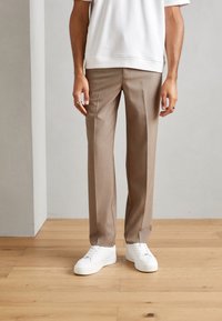 Beige skreddersydde bukser laget av et glatt stoff, kombinert med hvite sneakers. Bukser har en rett ben-design og subtile tværfold detaljer.
