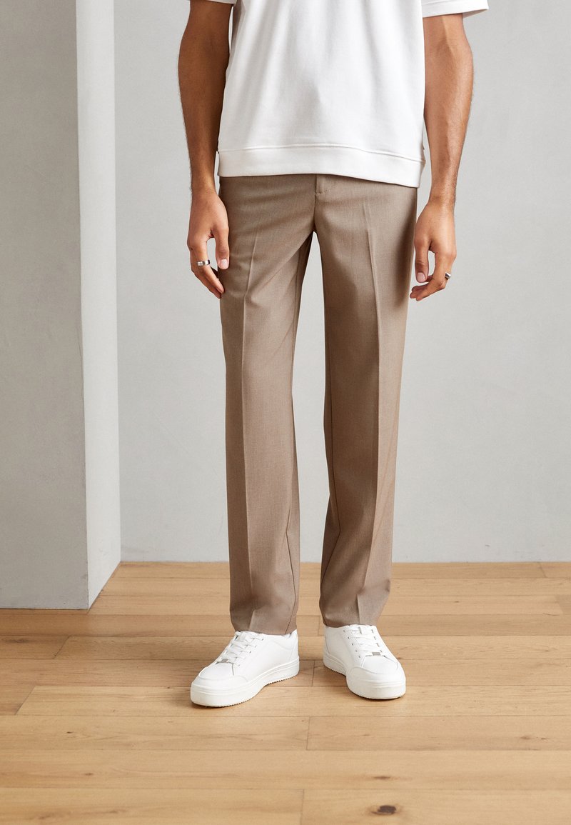Beige skreddersydde bukser laget av et glatt stoff, kombinert med hvite sneakers. Bukser har en rett ben-design og subtile tværfold detaljer.