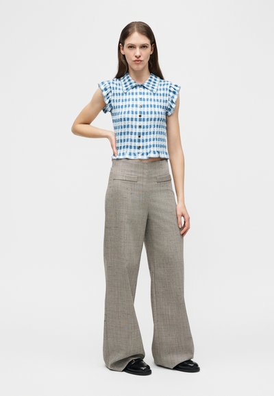 Γυναίκα που φοράει μπλε και λευκό καρό αμάνικο πουκάμισο με κουμπιά και φαρδιά γκρι παντελόνια με μαύρα παπούτσια, στέκεται με το ένα χέρι στη μέση.