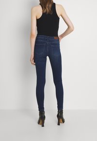 Skinny jeans in dark blue denim, with a high waist, back pockets, and a brown leather patch. Worn with black heeled boots.
