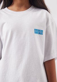 Osoba nosiaca biele tričko s modrým nápisom "AMERICAN VINTAGE" na ľavej strane hrude, zobrazená od krku po stred trupu.