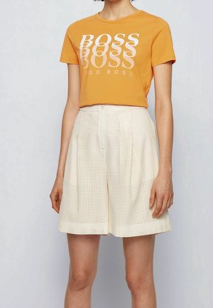 Vrouw draagt een oranje cropped T-shirt met de tekst "BOSS HUGO BOSS" en witte, hoog uitgesneden geperforeerde shorts met een rits aan de voorkant.