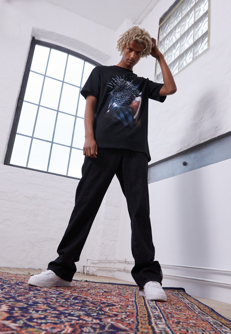 Black graphic t-shirt featuring a textured print of spikes, paired with black wide-leg jeans and white sneakers, set against a bright window.