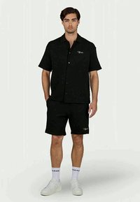 Camicetta nera a maniche corte con bottoni e taschino frontale, abbinata a pantaloni corti neri. Presenta il logo del marchio in bianco sui pantaloni e una texture liscia.