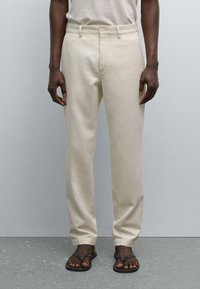 Pantalons beige clair avec une coupe droite, passants de ceinture et poches avant. Portés avec des tongs noires, mettant en valeur une texture tissée lisse.