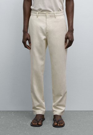 Light beige trousers with a straight fit, belt loops, and front pockets. Worn with black flip-flops, showcasing a smooth, woven texture.