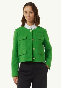 Groene textuurjasje met een ronde halslijn, gouden knopen en twee voorzakken, gedragen over een witte blouse met kraag.