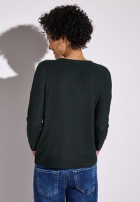 Mörkgrön, texturerad tröja med avslappnad passform och trekvartärmar, kombinerad med ljusblå denimjeans.