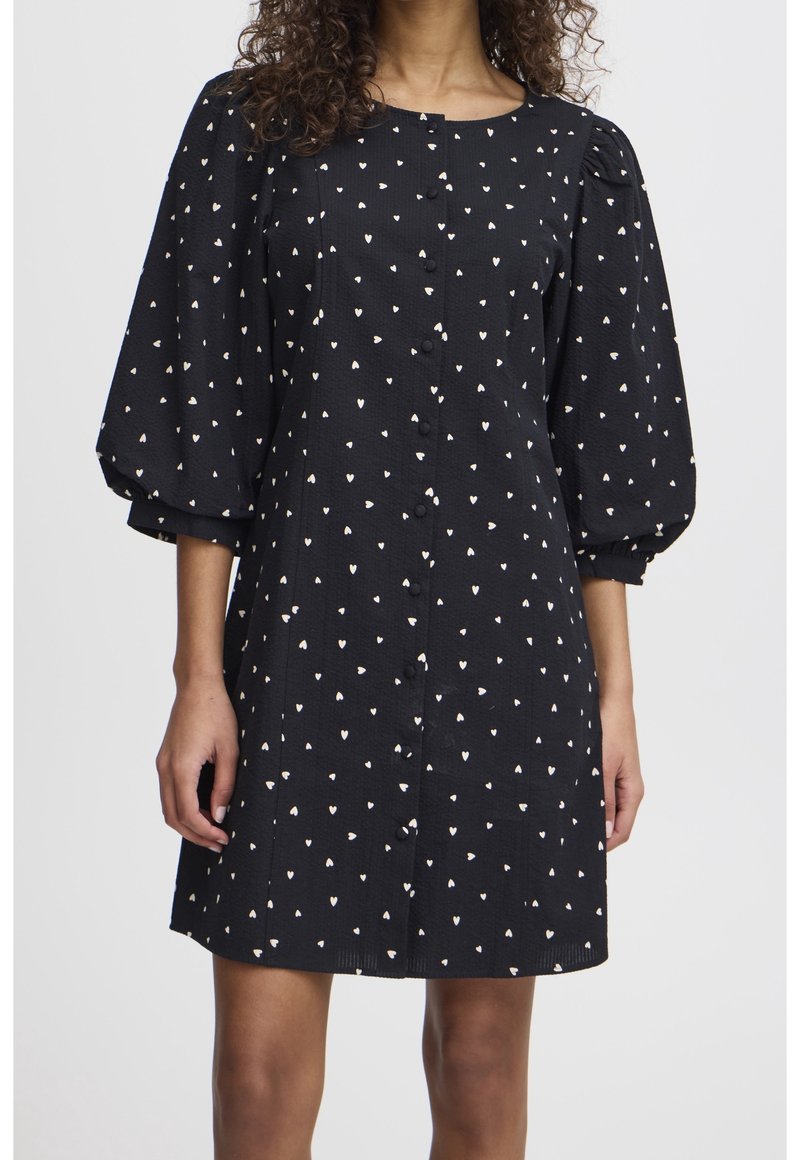 Femme portant une robe noire avec des motifs de cœurs blancs, des manches trois-quarts bouffantes et une patte de boutonnage à l'avant.