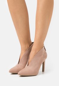 Zapatos de tacón alto de ante rosa con punta afilada, que presentan un diseño de corte profundo y un zipper trasero, expuestos sobre un fondo blanco.