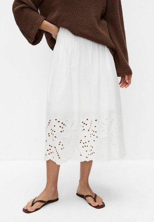 Personne porte une jupe midi blanche avec un ourlet à découpes florales, un pull en maille marron et des tongs marron, debout devant un fond blanc uni.
