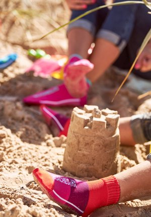 Calzini rosa e rossi colorati con un design a gradiente, indossati su piedi nella sabbia vicino a un castello di sabbia, che mostrano un ambiente giocoso da spiaggia.