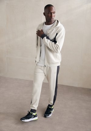 Sudadera beige con cremalleras y detalles negros, pantalones deportivos grises con una franja lateral, combinados con zapatos deportivos negros con detalles en verde neón.