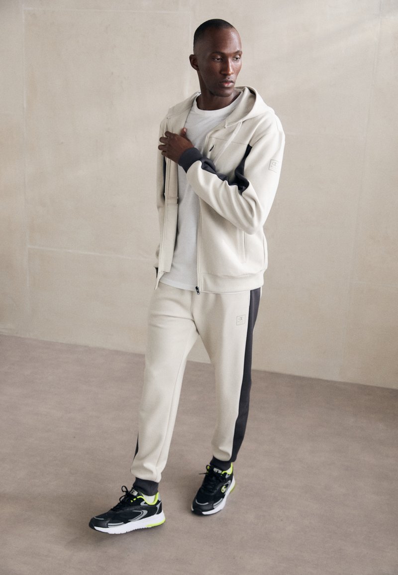 Sudadera beige con cremalleras y detalles negros, pantalones deportivos grises con una franja lateral, combinados con zapatos deportivos negros con detalles en verde neón.