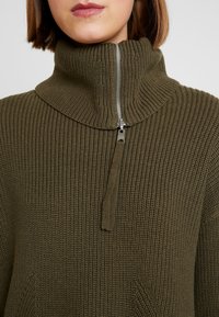 Maglione a coste di colore verde oliva, con colletto alto e mezza cerniera. Presenta un motivo testurizzato e una piccola tasca frontale.
