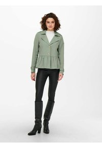 Giacca peplum verde chiaro con colletto, bottoni dorati e maniche lunghe, abbinata a pantaloni neri aderenti e stivali neri fino al ginocchio.