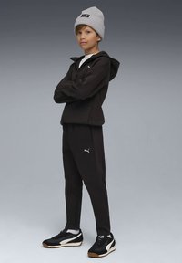 Jauns cilvēks melnā Puma sporta tērpā, pelēkā cepurē un melnās kedās stāv ar sakrustotām rokām uz pelēka fona.