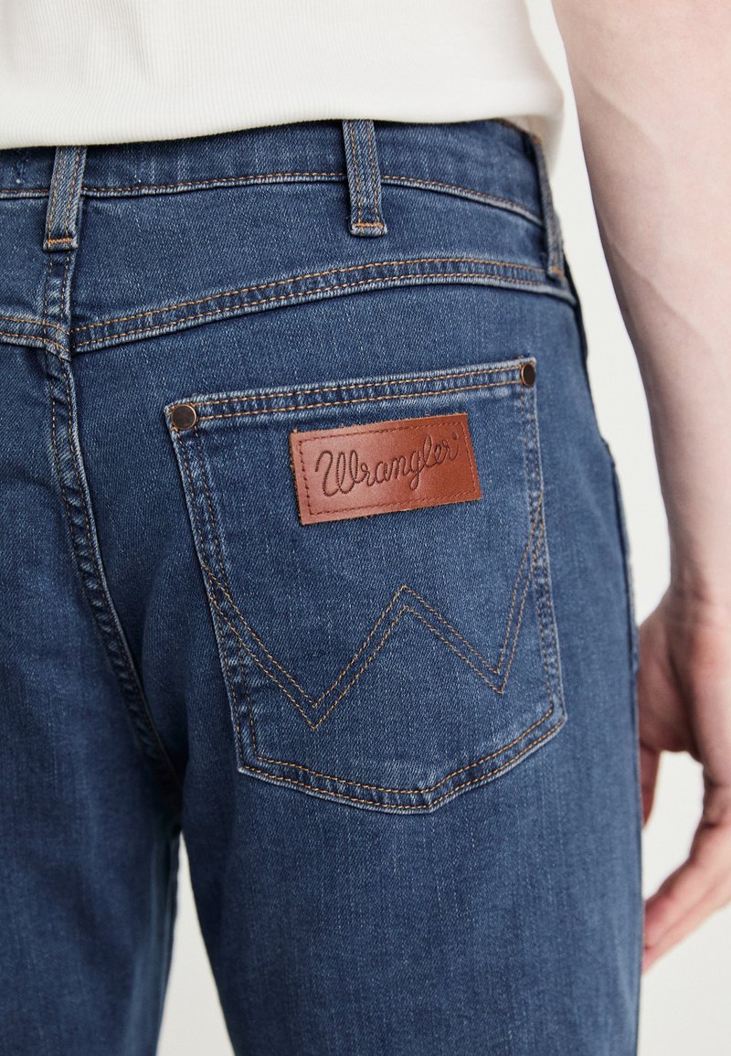 Niebieskie dżinsy z denimu z brązową skórzaną naszywką oznaczoną "Wrangler". Posiadają zygzakowaty ścieg na tylnej kieszeni oraz standardowy pas.