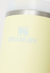 Stanley vakuumflaske, lett pastellgul, rustfritt stål bånd, jevn tekstur, har preget logo med et løvegrafikk.