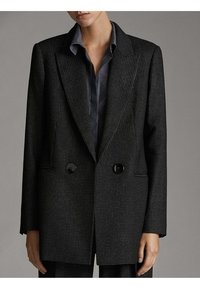 Femme portant un blazer croisé texturé gris foncé par-dessus une chemise boutonnée foncée, debout devant un fond gris neutre.