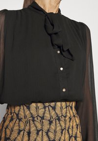 Blouse noire avec manches longues transparentes, ornée d'un nœud à la gorge et de boutons argentés. La jupe présente un motif de feuilles dorées sur un fond bleu marine.