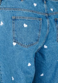 Zilā džinsa bikses ar baltām sirds formas apdrukām, ar aizmugures kabatām, klasiskām šuvēm un nedaudz nolietotu tekstūru.