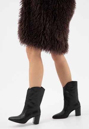 Beine in schwarzen kniehohen Lederstiefeln mit Absatz, kombiniert mit einem braunen, flauschigen Rock auf weißem Hintergrund.
