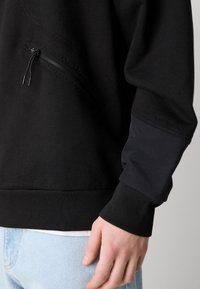 Primo piano di una persona che indossa una felpa nera con tasca con cerniera e toppa rinforzata sul gomito, abbinata a jeans azzurri.