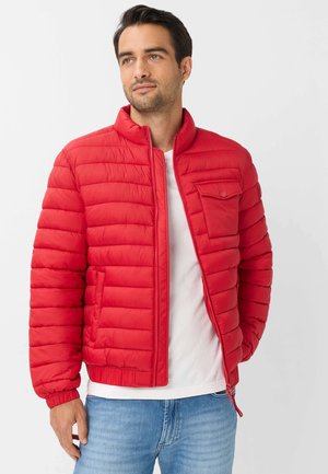 Mand iført rød quiltet jakke over hvid skjorte og blå jeans, stående med hånd i lommen, kigger let til siden.